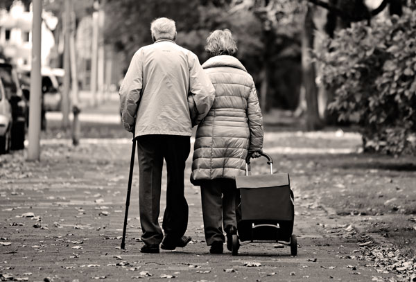 An elderly couple walking arm in arm are seen from the back, with the man on the left walking with a cane, while the woman on the right is dragging a wheeled grocery bag.